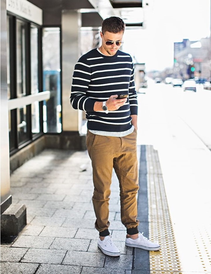 homem branco em calçada estando em pé e olhando seu celular. Ele está vestindo óculos, suéter, e um dos estilos de calça masculina mais comuns atualmente, a jogger