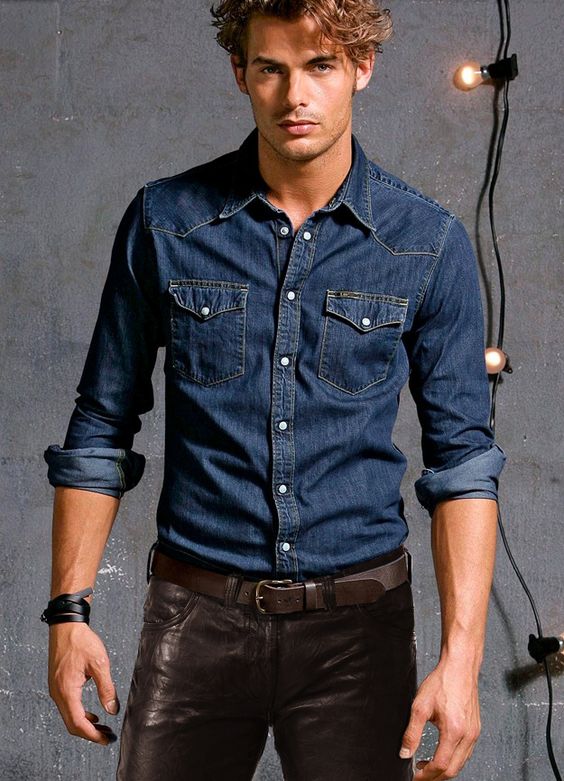 Homem de louro posando para foto com camisa jeans de mais escura e calça de couro marrom.