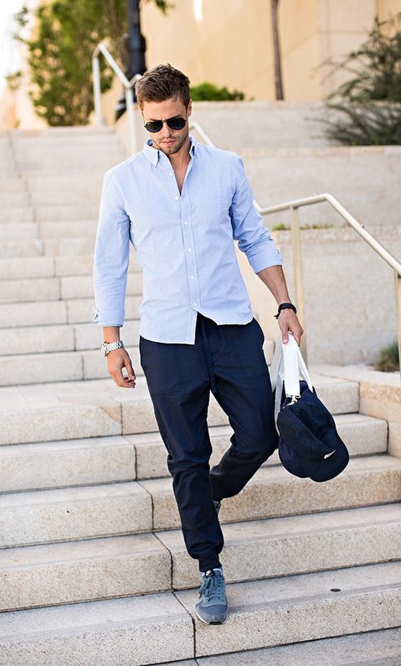 Foto de corpo inteiro de homem branco com camisa social clara, segurando uma bolsa e vestindo uma calça jogger azul marinho e tênis Street.