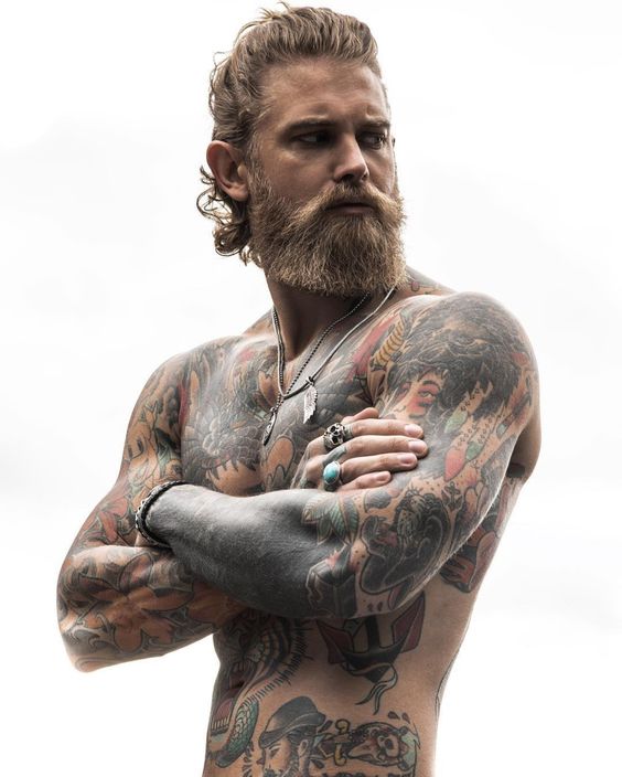 Homem descamisado de braços cruzados, todo tatuagem e com barba estilo rustica de tamanho médio.