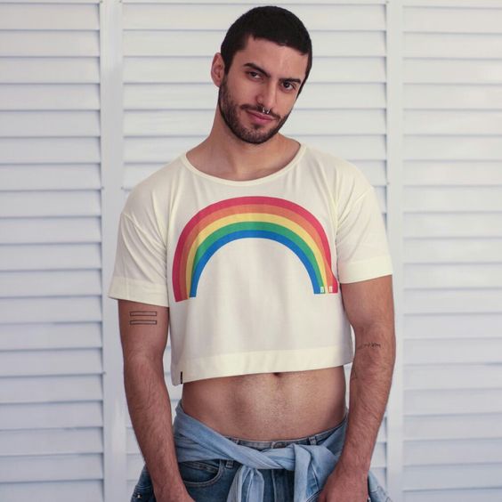Rapaz branco fazendo pose para foto com uma camisa  branco com um arco-iris colorido em homenagem aos LGBTQIA+