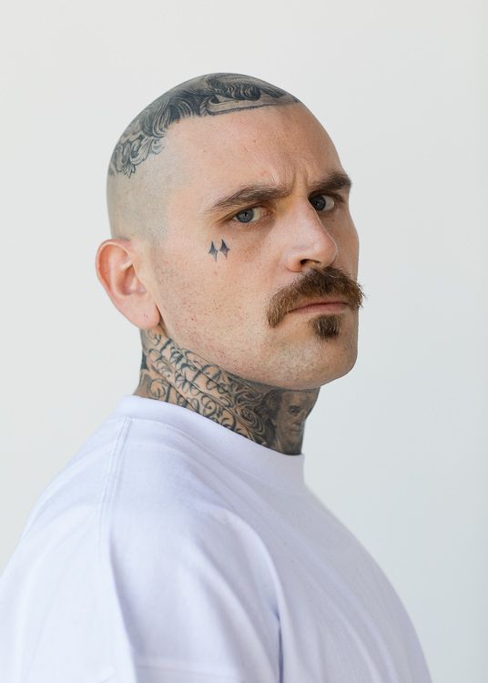 Homem branco com rosto tatuagem e bigode.