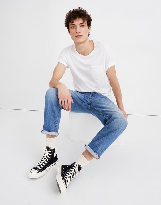 Rapaz jovem branco sentado com camisa branca e calça jeans reta azul padrão.