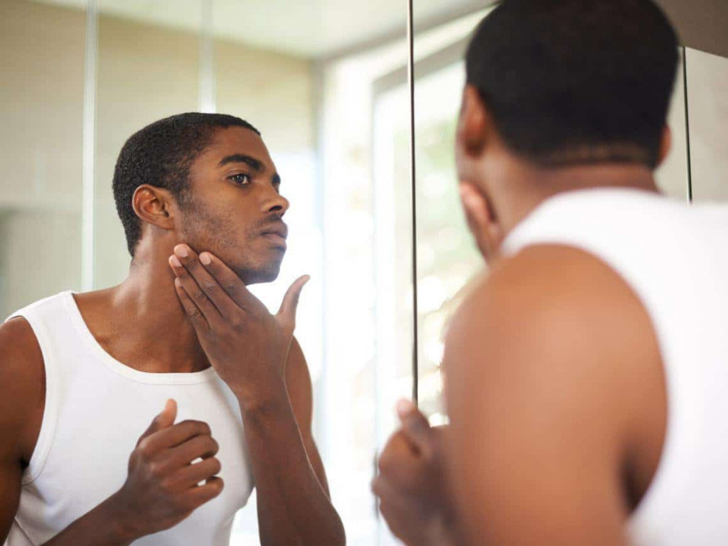 Homem negro observando seu rosto com foliculite no espelho